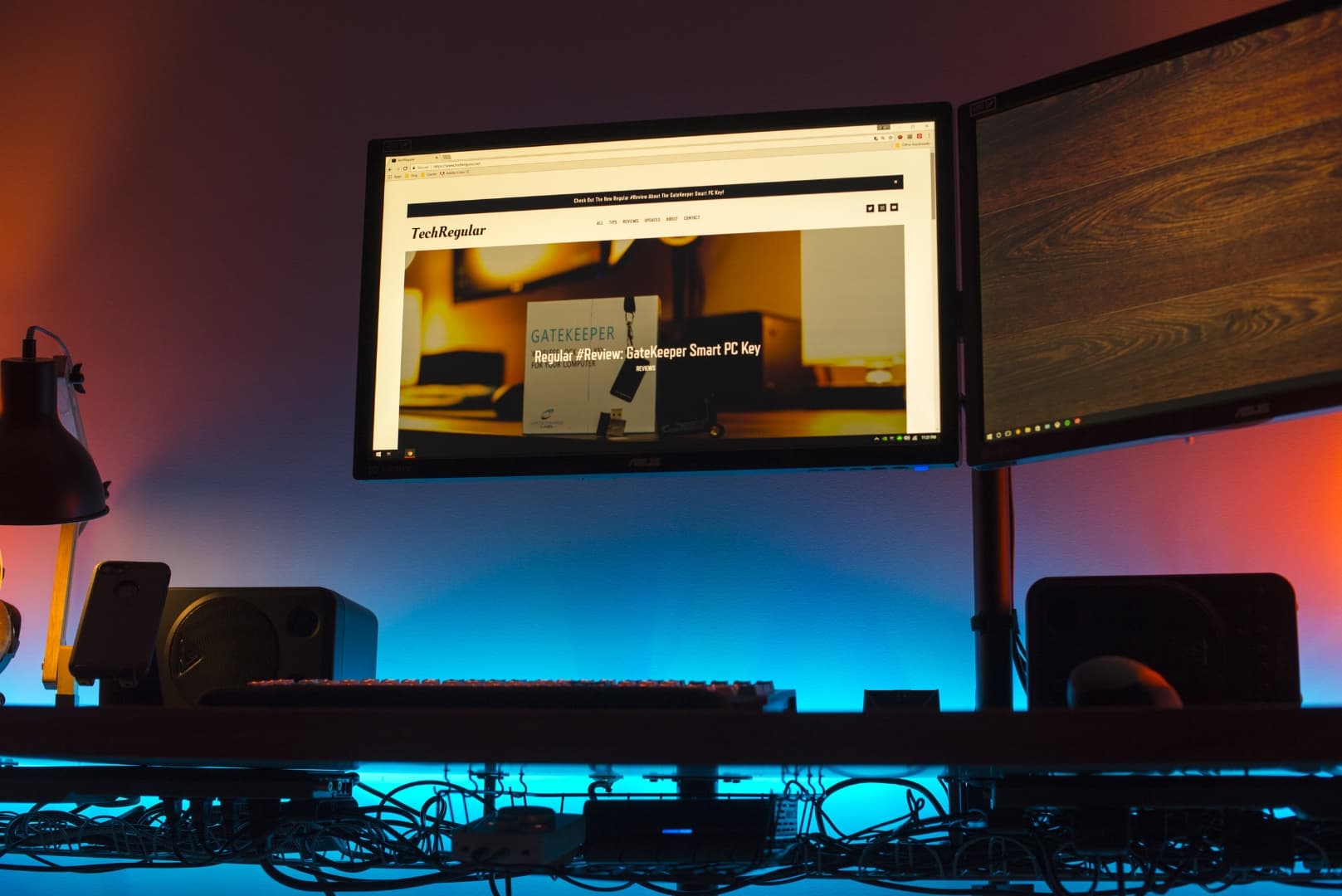 Monitors mounted on a desk with a monitor arm with colored lights against wall behind the monitors